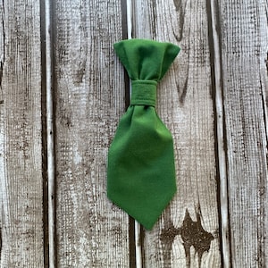 May include: A dark green fabric bow tie with a loop closure. The bow tie is photographed against a wood background.