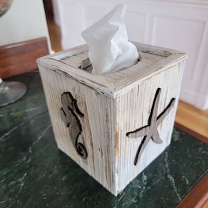 May include: A square, wooden tissue box with a distressed, whitewashed finish. The box features cut-out designs of a seahorse and a starfish. White tissues are visible, emerging from the top opening. The box is on a green marble surface.