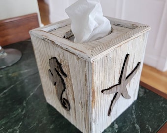 May include: A square, wooden tissue box with a distressed, whitewashed finish. The box features cut-out designs of a seahorse and a starfish. White tissues are visible, emerging from the top opening. The box is on a green marble surface.