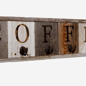 May include: Wooden wall-mounted coat rack with five hooks. The rack is painted in shades of brown, white, and gray. The word "COFFEE" is spelled out in black letters on the front of the rack.