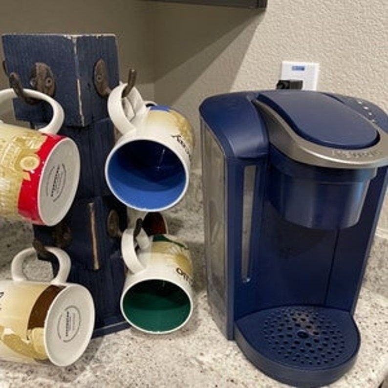 Op de afbeelding: Een blauwe koffiezetapparaat met een zilveren bovenkant en een houten bekerrek met vier bekers die eraan hangen. De bekers zijn versierd met verschillende ontwerpen.
