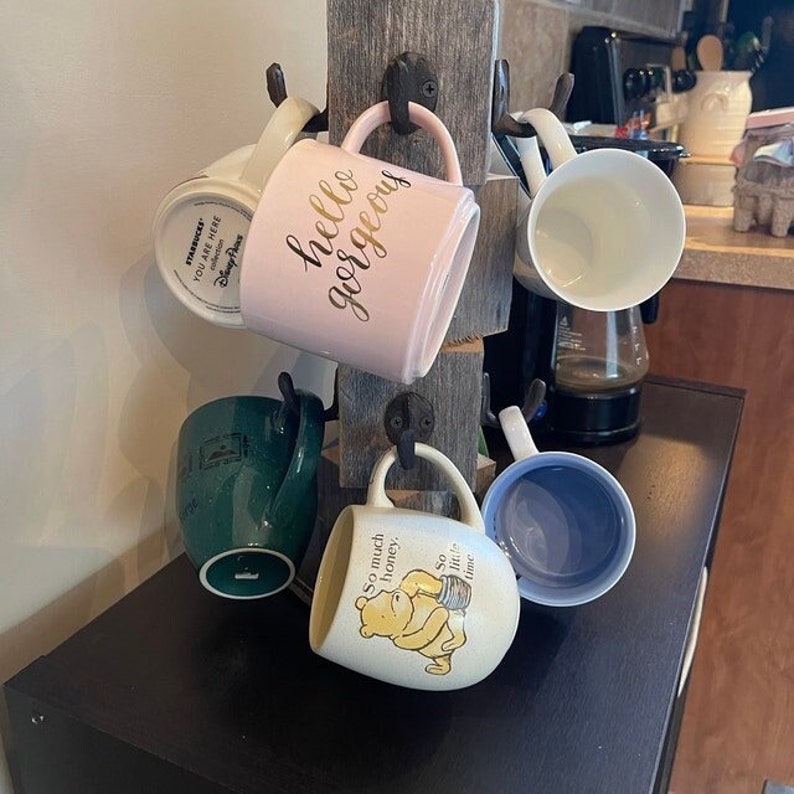 Op de afbeelding: Een houten bekerrek met vijf bekers die eraan hangen. De bekers zijn in verschillende kleuren en ontwerpen, waaronder een roze beker met gouden letters die "hello gorgeous" zeggen, een groene beker met een ontwerp, een witte beker met een Winnie the Pooh-ontwerp en een witte beker met gouden letters die "Starbucks You Are Here Disney Parks" zeggen.