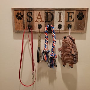 May include: A wooden pet leash and toy holder with the name "SADIE" in brown letters. The holder has paw print designs and includes a red leash, a brush, a rope toy, and a stuffed hedgehog toy.