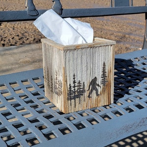 May include: A rustic wooden tissue box holder with a silhouette of Bigfoot walking through a forest. The box is sitting on a gray metal bench.