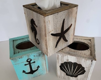 May include: Three wooden tissue box covers with nautical themes. The top cover is whitewashed wood with a brown top and a seahorse, starfish, and seashell design. The bottom left cover is painted turquoise blue with an anchor design. The bottom right cover is whitewashed wood with a seashell design.
