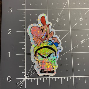 Puede incluir: Una pegatina holográfica con los personajes de dibujos animados Grim, Billy y Mandy. Grim está encima, con Billy y Mandy debajo. La pegatina tiene un acabado brillante e iridiscente, que refleja varios colores.