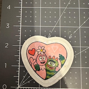 May include: Heart-shaped sticker featuring two cartoon characters in pink and green, with a pink background and a red heart. The sticker has a white border and is on a black surface with measurement markings.