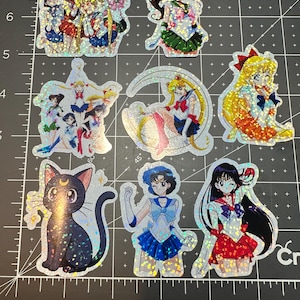 以下が含まれることがあります： 様々なポーズの、アニメキャラクターが描かれたホログラフィックステッカーのコレクション。ステッカーには、黒猫や三日月模様など、鮮やかな髪と衣装のキャラクターが描かれています。ステッカーは黒とグレーのグリッド上にあります。