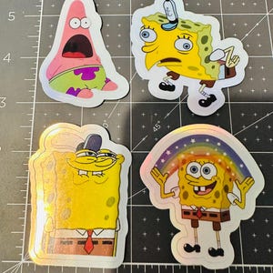 Puede incluir: Cuatro pegatinas holográficas con personajes de una popular serie animada. Una pegatina representa una estrella de mar rosa con la boca abierta. Tres pegatinas muestran un personaje de esponja amarilla en diferentes poses, incluida una con un arcoíris.