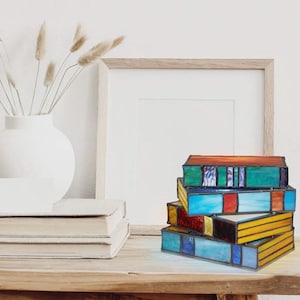 May include: A stack of stained glass book-shaped lamps with a blue, green, yellow, and red color scheme. The lamps are on a wooden table with a vase of dried flowers and a picture frame in the background.