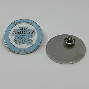 May include: A round, light blue and silver pin with the text "Zeta Amicae" and "1948". The pin features a logo with two hands clasped together. The pin is made of metal and has a pin back.