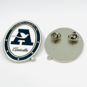 May include: A white oval pin with a blue border and a white letter A with a white bird flying above it. The text "Zeta Phi Beta Sorority, Inc." is around the top of the oval and the text "Amicette" is below the letter A. The pin has a silver back with a pin and a clasp.