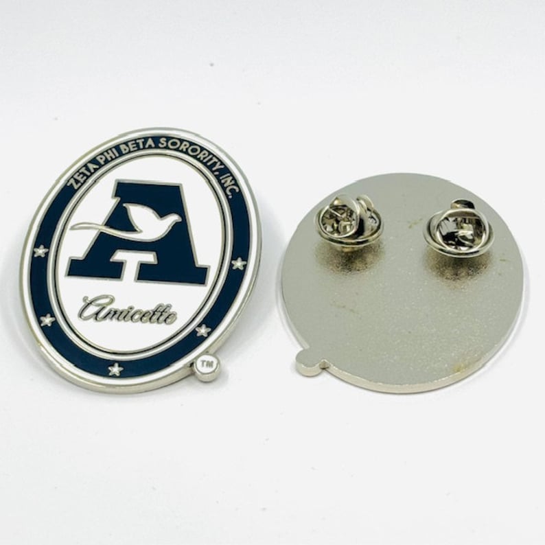 May include: A white oval pin with a blue border and a white letter A with a white bird flying above it. The text "Zeta Phi Beta Sorority, Inc." is around the top of the oval and the text "Amicette" is below the letter A. The pin has a silver back with a pin and a clasp.