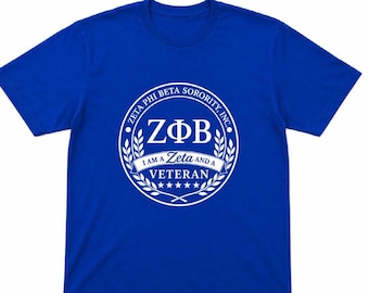 Op de afbeelding: Een koningsblauw T-shirt met korte mouwen en een witte cirkelvormige afbeelding op de borst. Het ontwerp bevat de tekst ZETA PHI BETA SORORITY, INC., de Griekse letters ZΦB en de zin I AM A ZETA AND A VETERAN met vijf kleine sterren.