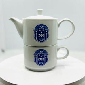 May include: A white ceramic tea set with a blue and white logo that says "ZOB" on the side. The set includes a teapot and a cup.