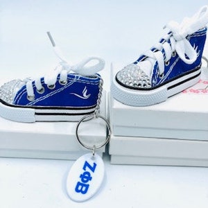 Könnte beinhalten: Blaue und weiße Canvas-Sneaker mit Strasssteinen, befestigt an einem Schlüsselring mit einem weißen ovalen Anhänger, auf dem "Zo&B" steht