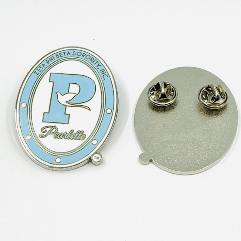 May include: A blue and white enamel pin with the letter "P" and the words "Zeta Phi Beta Sorority, Inc." and "Pearlette" in gold lettering. The back of the pin is silver with a safety pin.