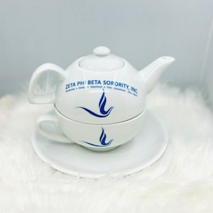 Puede incluir: Juego de té de cerámica blanca con tetera y taza. La tetera y la taza están apiladas una encima de la otra. La tetera tiene un logotipo azul que dice "Zeta Phi Beta Sorority, Inc." con las palabras "Scholarship, Service, Sisterhood, Finer Womanhood, Since 1920" debajo del logotipo. La taza tiene un logotipo azul de un pájaro con las alas extendidas.
