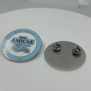 May include: A silver pin with a blue and white enamel design. The pin features the text "Zeta Amicae" and "Zeta Amicae" with a trademark symbol. The year "1948" is printed below the design.