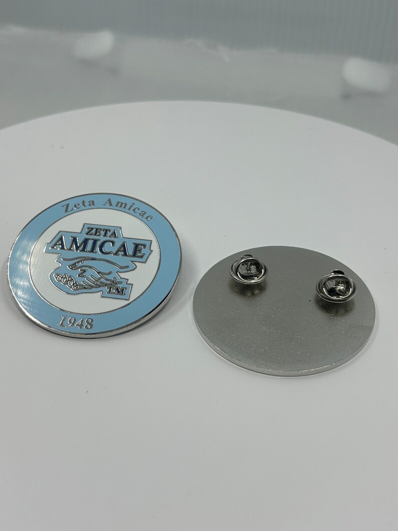 May include: A silver pin with a blue and white enamel design. The pin features the text "Zeta Amicae" and "Zeta Amicae" with a trademark symbol. The year "1948" is printed below the design.