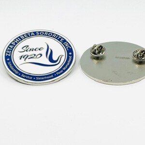 May include: A white and blue enamel pin with the text "Zeta Phi Beta Sorority, Inc." and "Since 1920" in a blue circle. The pin also features a blue wave and a blue flame. The text "Scholarship, Service, Sisterhood, Finer Womanhood" is written around the circle.