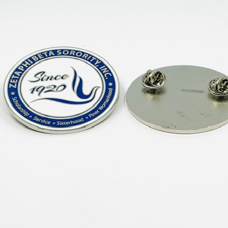 May include: A white and blue enamel pin with the text "Zeta Phi Beta Sorority, Inc." and "Since 1920" in a blue circle. The pin also features a blue wave and a blue flame. The text "Scholarship, Service, Sisterhood, Finer Womanhood" is written around the circle.