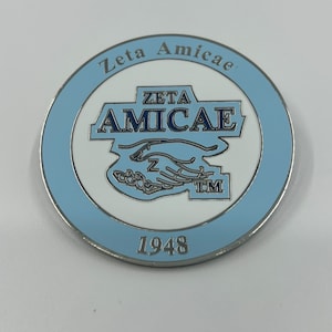 May include: A silver-colored coin with a blue and white design. The coin features the text "Zeta Amicae" and "Zeta Amicae TM" in a white circle with a blue border. The year "1948" is printed below the circle.