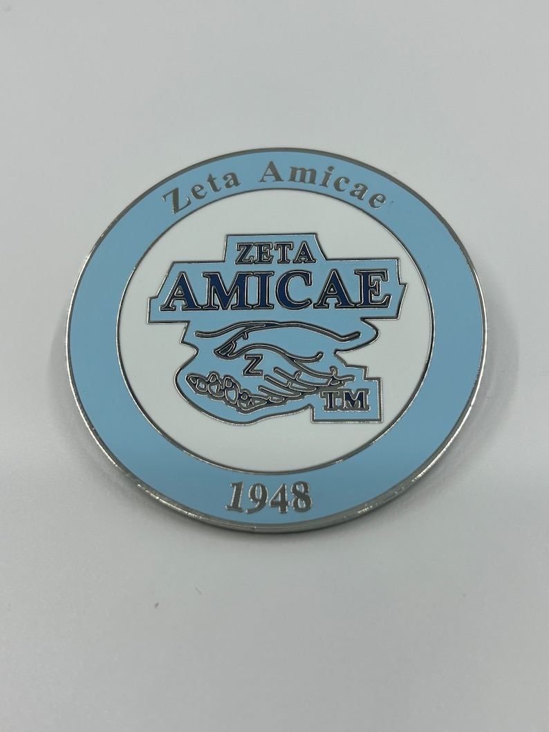 May include: A silver-colored coin with a blue and white design. The coin features the text "Zeta Amicae" and "Zeta Amicae TM" in a white circle with a blue border. The year "1948" is printed below the circle.
