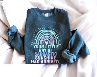 May include: A dark gray sweatshirt with a rainbow graphic and the text "Your little ray of sarcastic sunshine has arrived."