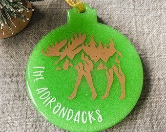 May include: A green Christmas ornament with a gold moose silhouette and the text "THE ADIRONDACKS" in white.