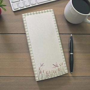 May include: A rectangular notepad with a green and white gingham border. The bottom of the notepad features a watercolor illustration of two ducks flying over reeds. A black pen, a white mug, and a keyboard are also visible.