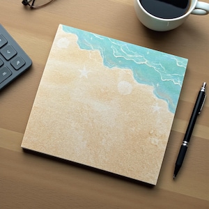 May include: A square notepad with a beach scene design. The top right corner features a turquoise ocean wave design, with a sandy beach below. A black pen and a cup of coffee are next to the notepad.