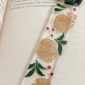 May include: A clear bookmark with a floral design. The bookmark features three stylized flowers in shades of brown and yellow, with green leaves and red dots scattered throughout.