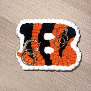 May include: A crocheted orange and black tiger stripe design with a white border. The design is shaped like the letter "B". There are gold hoop earrings and rings on top of the design.