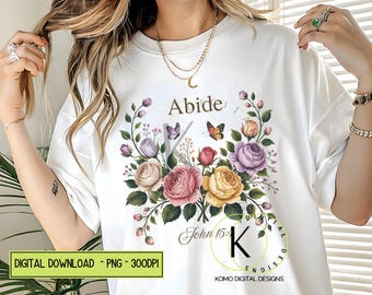 May include: White t-shirt featuring a floral design with roses in shades of pink, yellow, and purple, along with butterflies. The word "Abide" is above the floral arrangement, with "John 15:" below. The shirt has a round neckline.