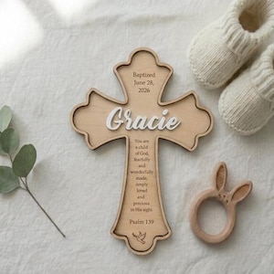 May include: A light-coloured wooden cross featuring the name Gracie in white script, the text Baptised June 28, 2026, a religious verse from Psalm 139, and a small dove icon. The cross is accompanied by a pair of cream-coloured knit baby booties.