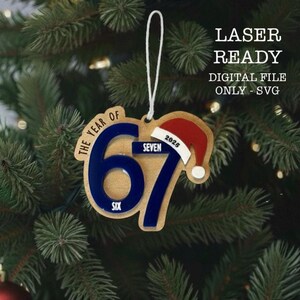 Peut inclure: Ornement de Noël en bois avec le texte "THE YEAR OF 67" en bleu et "SIX" et "SEVEN" en blanc. Un bonnet de Père Noël avec l'année "2025" est également inclus. L'ornement est suspendu à un sapin de Noël. Le texte "LASER READY DIGITAL FILE ONLY - SVG" est également inclus.
