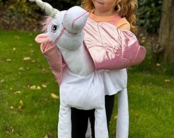 May include: A child wearing a pink and white unicorn costume. The costume features a plush unicorn head, pink wings, and long white legs with pink hooves.