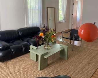 May include: A living room with a black leather sectional sofa, a green coffee table, and a brown woven rug. There is a large orange sphere hanging from the ceiling and a black leather chair in the background.