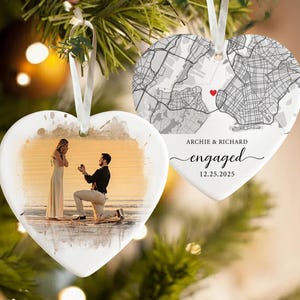 May include: Heart-shaped ceramic ornament with a photo of a proposal on one side and a map with a red heart on the other. The ornament has a white ribbon for hanging. Text on the ornament reads "Archie & Richard engaged 12.25.2025."
