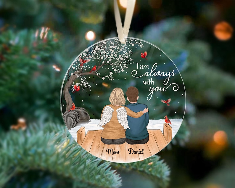 Puede incluir: Adorno navide&ntilde;o redondo y transparente con un dise&ntilde;o impreso. Presenta a una pareja sentada en un muelle de madera, con las palabras "I am always with you". Tambi&eacute;n se representan cardenales y un &aacute;rbol. Los nombres "Mom" y "Daniel" est&aacute;n impresos debajo.