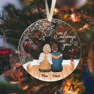 Puede incluir: Adorno navide&ntilde;o redondo y transparente con la frase "I am always with you". El adorno presenta a una pareja sentada en un muelle de madera, con una persona con alas de &aacute;ngel. Los nombres "Mom" y "Daniel" est&aacute;n escritos debajo.