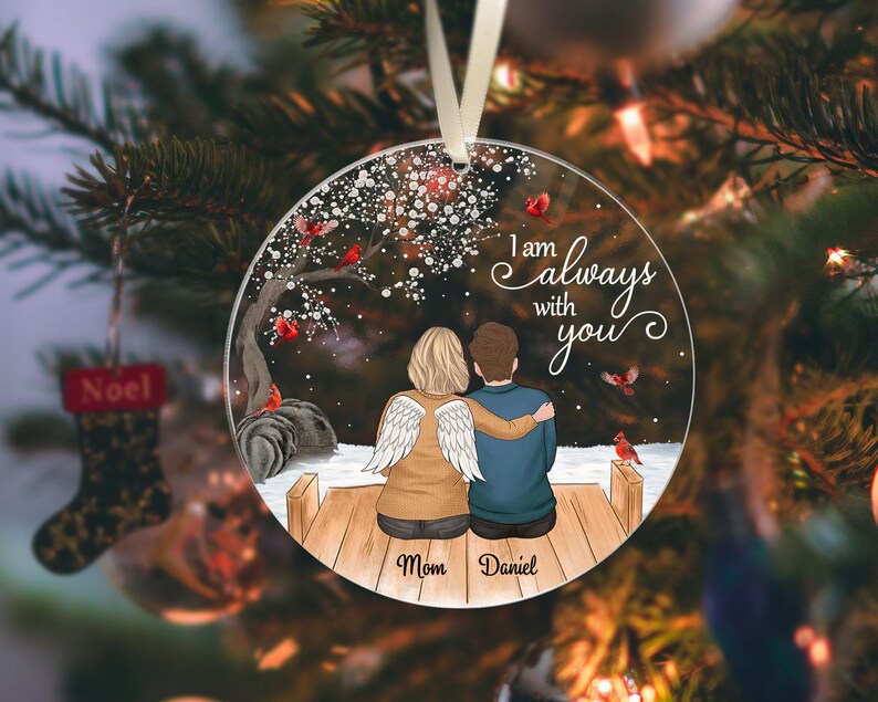 Puede incluir: Adorno navide&ntilde;o redondo y transparente con la frase "I am always with you". El adorno presenta a una pareja sentada en un muelle de madera, con una persona con alas de &aacute;ngel. Los nombres "Mom" y "Daniel" est&aacute;n escritos debajo.