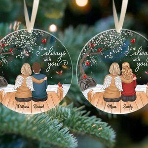 Puede incluir: Dos adornos navide&ntilde;os redondos y transparentes con un dise&ntilde;o impreso. Cada adorno presenta a una pareja sentada en un muelle de madera bajo un &aacute;rbol con p&aacute;jaros rojos. El texto "I am always with you" est&aacute; escrito en cada adorno. Un adorno dice "Patricia Daniel" y el otro dice "Mom Emily".