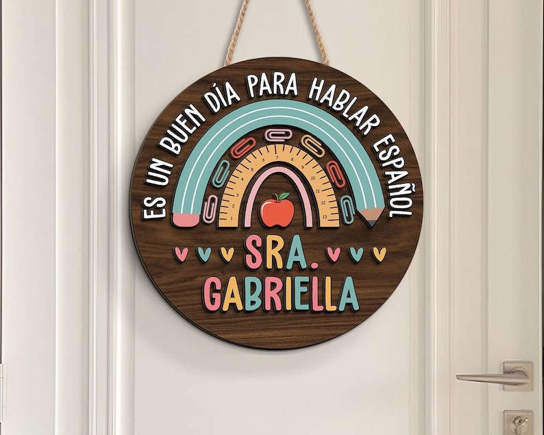 May include: A wooden round sign with a rainbow design and the text "Es un buen d&iacute;a para hablar espa&ntilde;ol Sra. Gabriella". The rainbow is made of blue, pink, and yellow paper clips and a ruler. There is a red apple in the center of the rainbow.