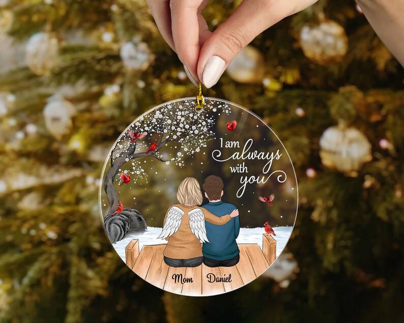 Puede incluir: Un adorno navide&ntilde;o redondo y transparente con un colgador dorado. El adorno presenta una pareja con alas de &aacute;ngel, un &aacute;rbol y el texto "I am always with you". Tambi&eacute;n se incluyen los nombres "Mom" y "Daniel".