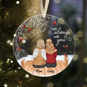 Puede incluir: Adorno redondo y transparente con una conmovedora escena de dos figuras abraz&aacute;ndose en un muelle de madera, con el texto "I am always with you". Una figura tiene alas de &aacute;ngel. Tambi&eacute;n se representan cardenales y un &aacute;rbol floreciente.