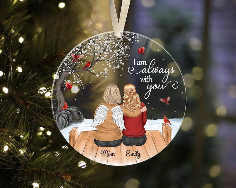 Puede incluir: Adorno redondo y transparente con una conmovedora escena de dos figuras abraz&aacute;ndose en un muelle de madera, con el texto "I am always with you". Una figura tiene alas de &aacute;ngel. Tambi&eacute;n se representan cardenales y un &aacute;rbol floreciente.
