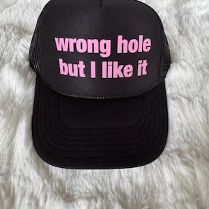 May include: Black trucker hat with a pink mesh back. The hat has the text "wrong hole but I like it" printed on the front in pink.