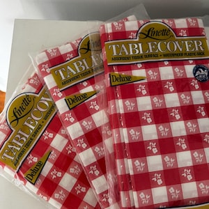 May include: Several packaged red and white checkered tablecloths. Each package features the word "TABLECOVER" and "Deluxe" in gold lettering. The tablecloths have a floral pattern and measure 54 inches by 108 inches.